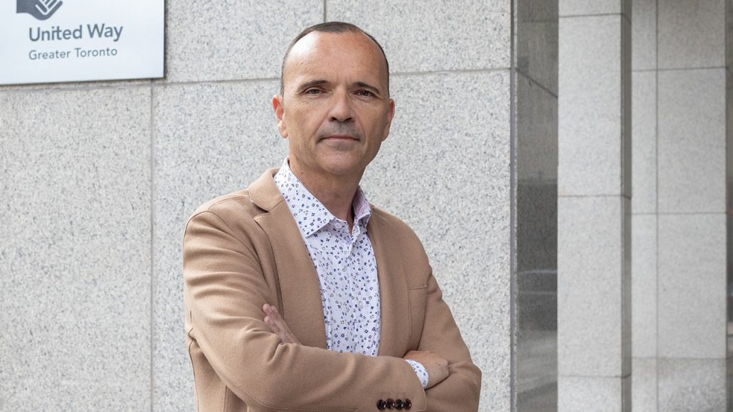 Daniele Zanotti, President and CEO of United Way Greater Toronto, stands in front of a gray wall. He is wearing a white shirt with blue dots and a tan jacket.