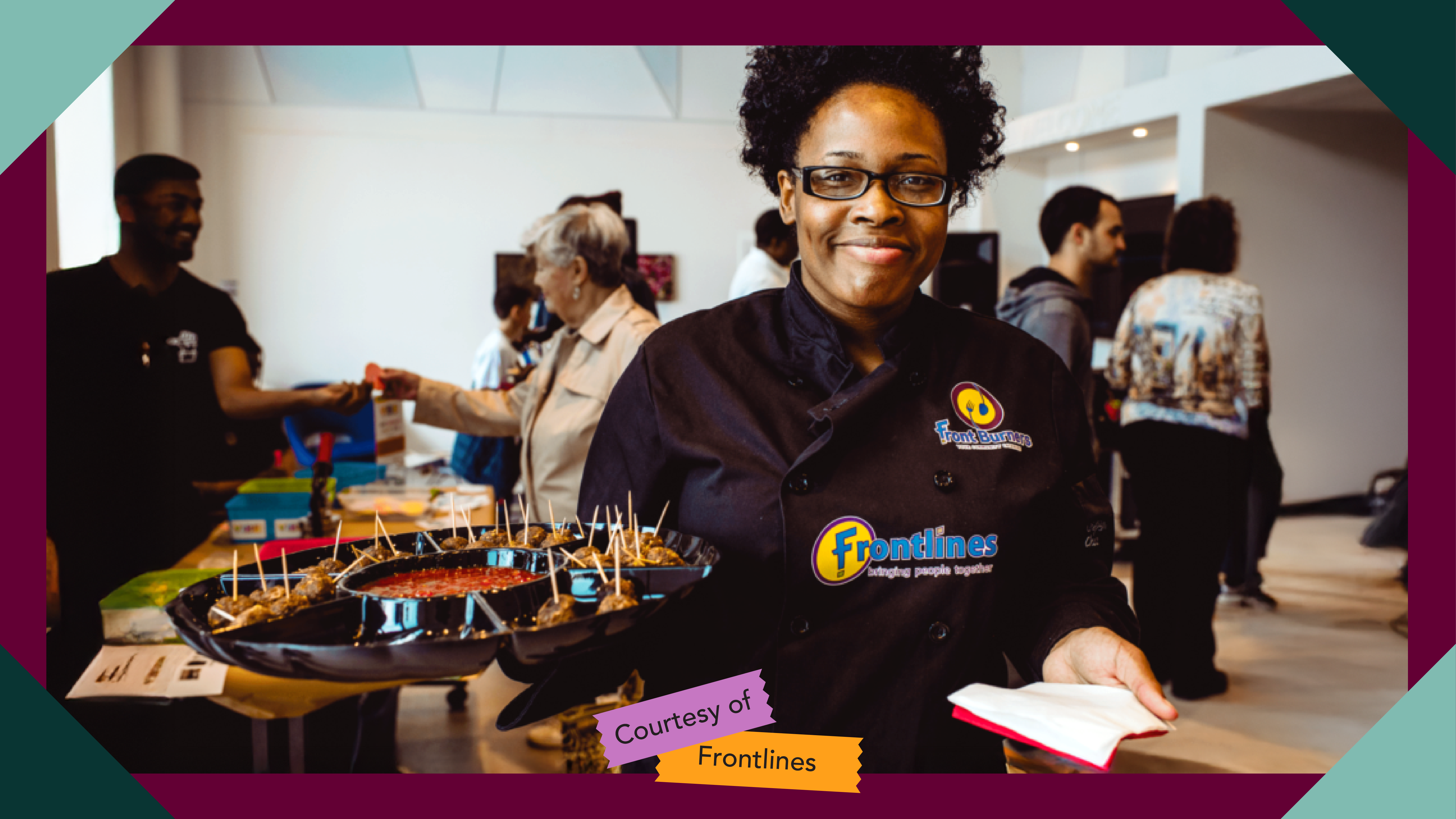 A person in a black Frontburners Catering shirt holding up a tray of food, with people in the background trying samples of food.