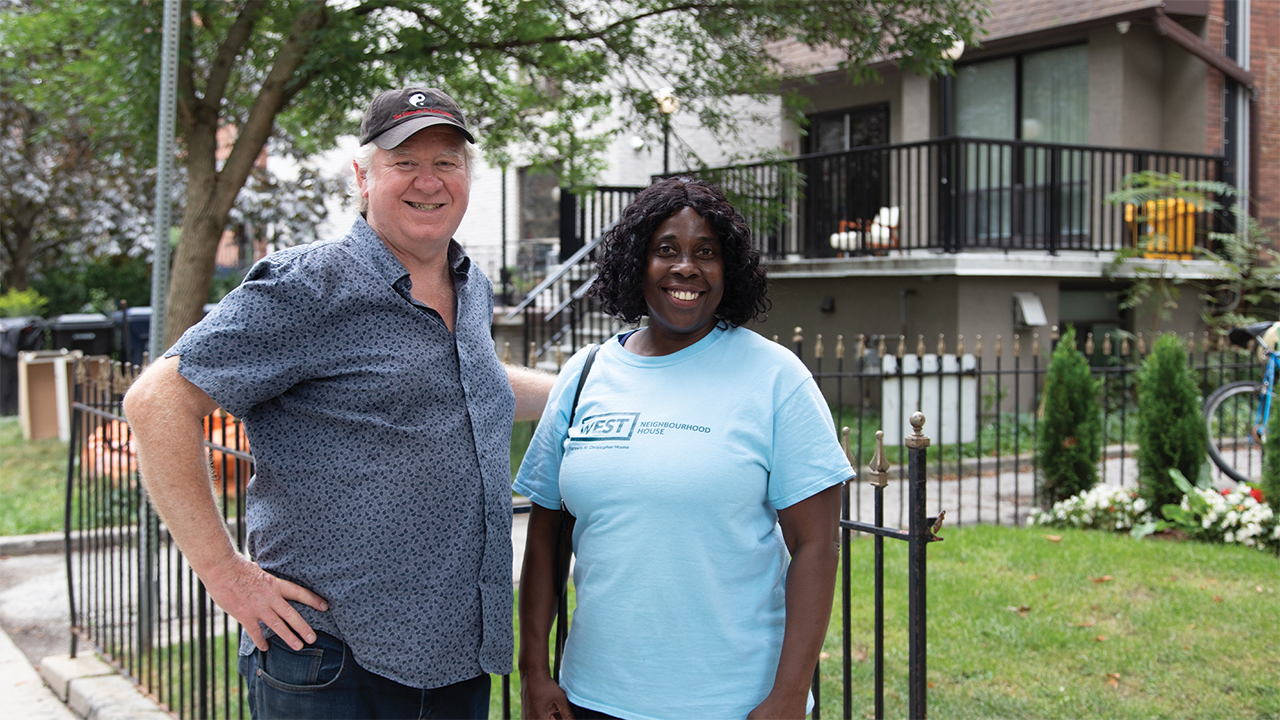 Victor, wearing a black baseball cap and a blue and black shirt, and Beryl-Ann, wearing a light blue shirt with 'West Neighbourhood House' written on it, smile as they pose next to each other.