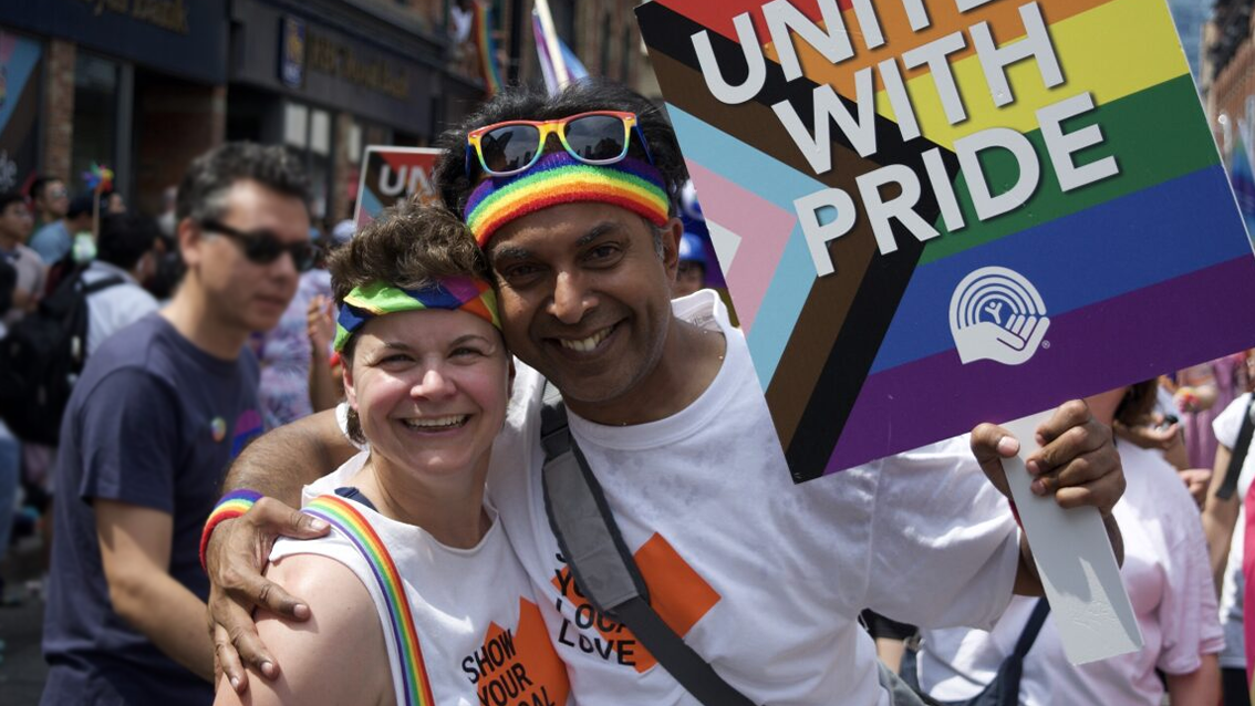People smiling and celebrating, wearing rainbow headbands and holding a sign that says ‘United with Pride’