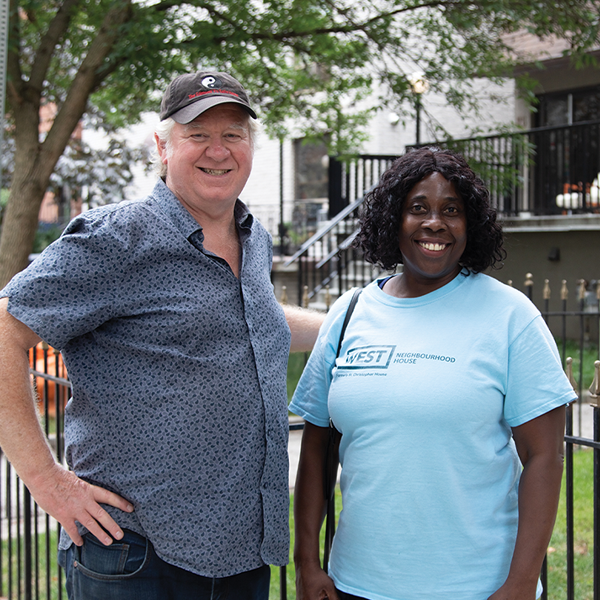 A picture of a white man and black woman outside smiling at the camera in front of neigbourhood.