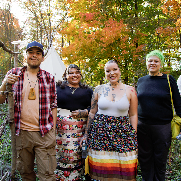 A group of Indigenous youth from ENAGB Indigenous Youth Agency stand in a wooded area and smile at the camera.