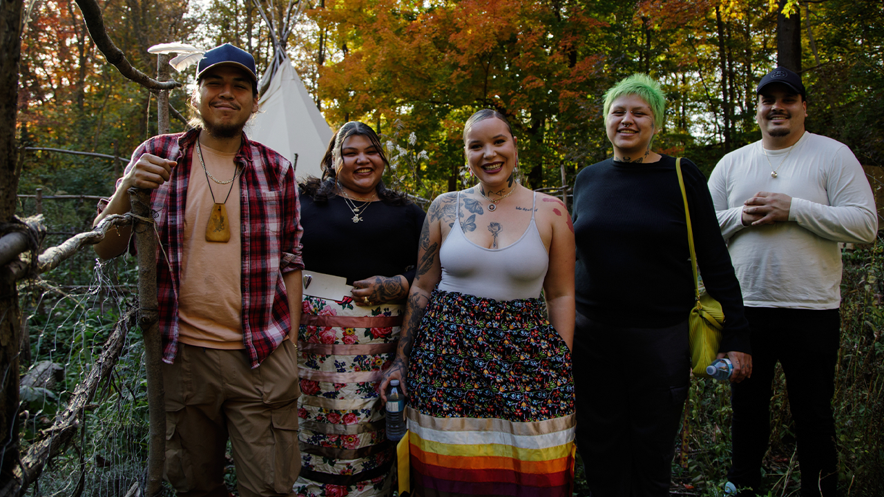 A group of Indigenous youth from ENAGB Indigenous Youth Agency stand in a wooded area and smile at the camera. 