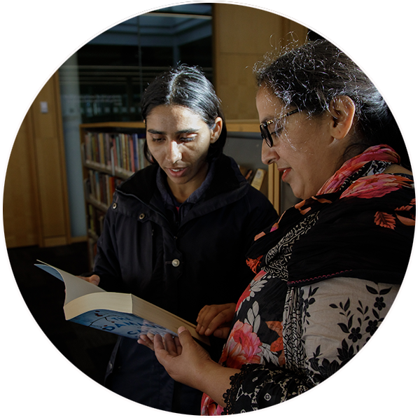 Two women, Muffad and Shenawar, read a book together in a library.