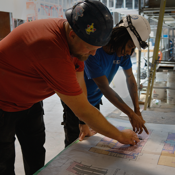 Two men wearing hard hats and leaning over to look at blueprints. 