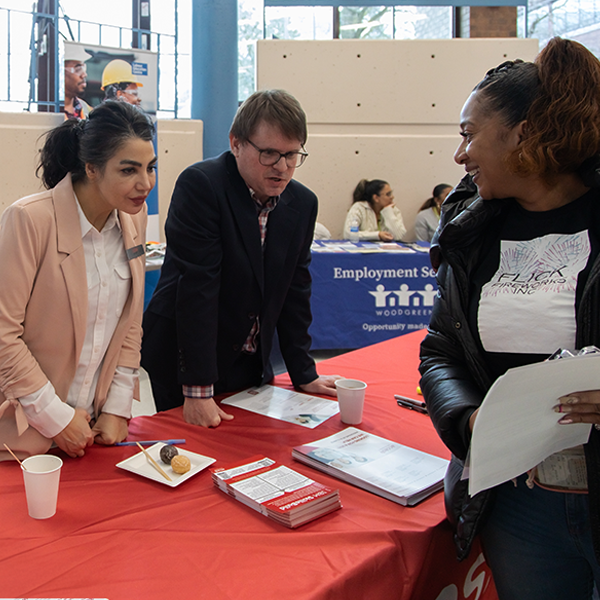 A woman stands talking to two people behind a booth at an employment fair.