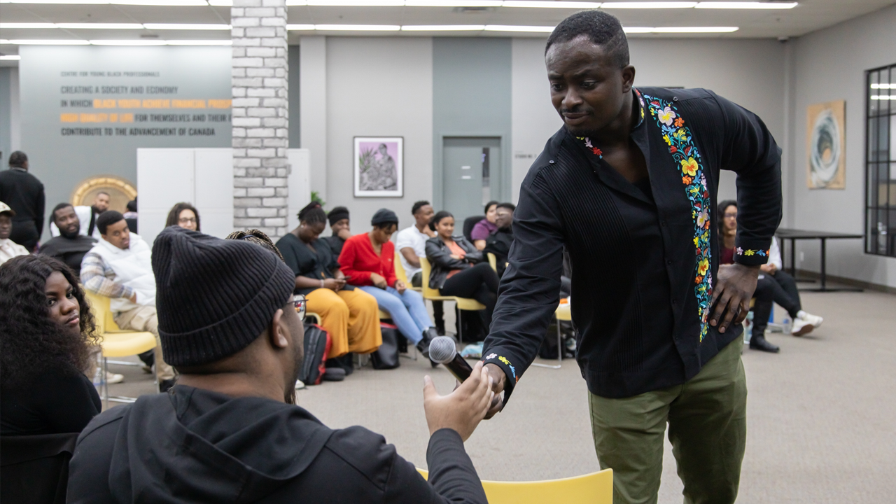 A Black man handing a microphone to another Black man sitting in the audience at an event at CEE Centre for Black Excellence.