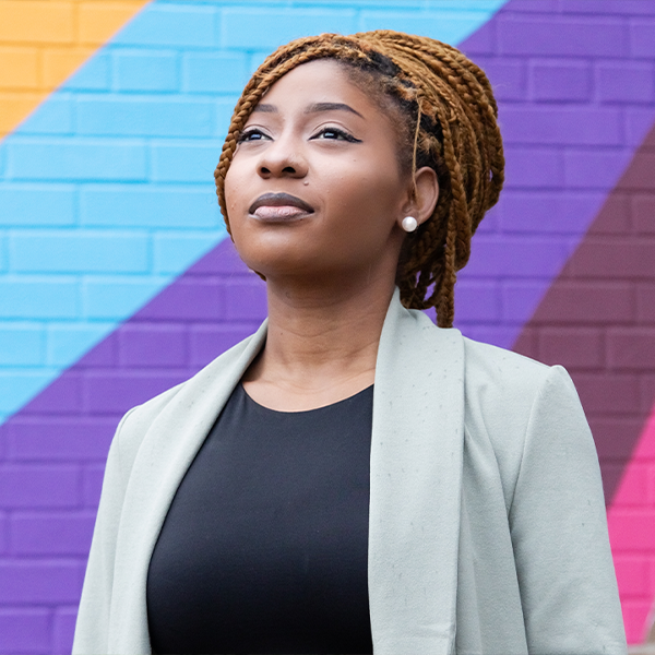 Demica, a Black woman, stands looking up and into the distance. She wears a black shirt and grey coat and is standing in front of a colourful mural.