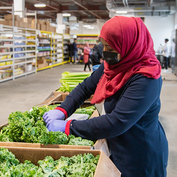 A woman wearing a face mask, red hijab and black shirt sorts through boxes of fresh produce.