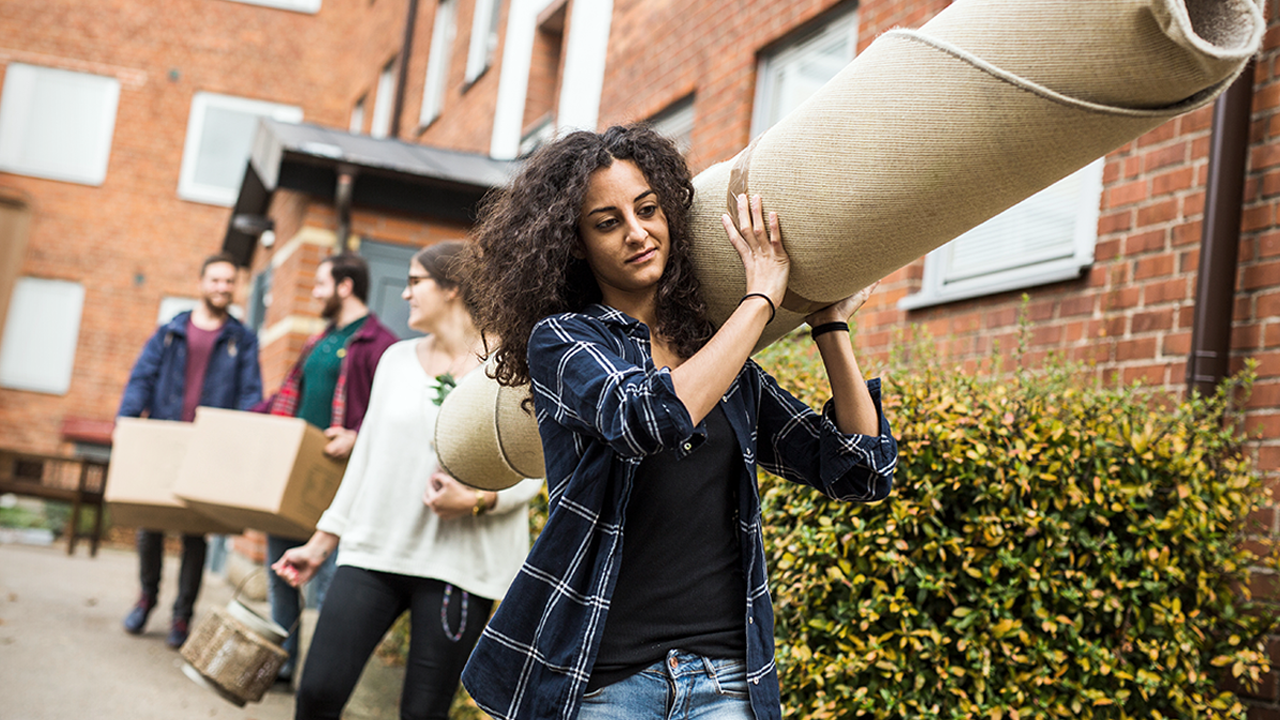 A women in foreground holding a rug, and three other people in the background holding boxes, showing people help someone move.